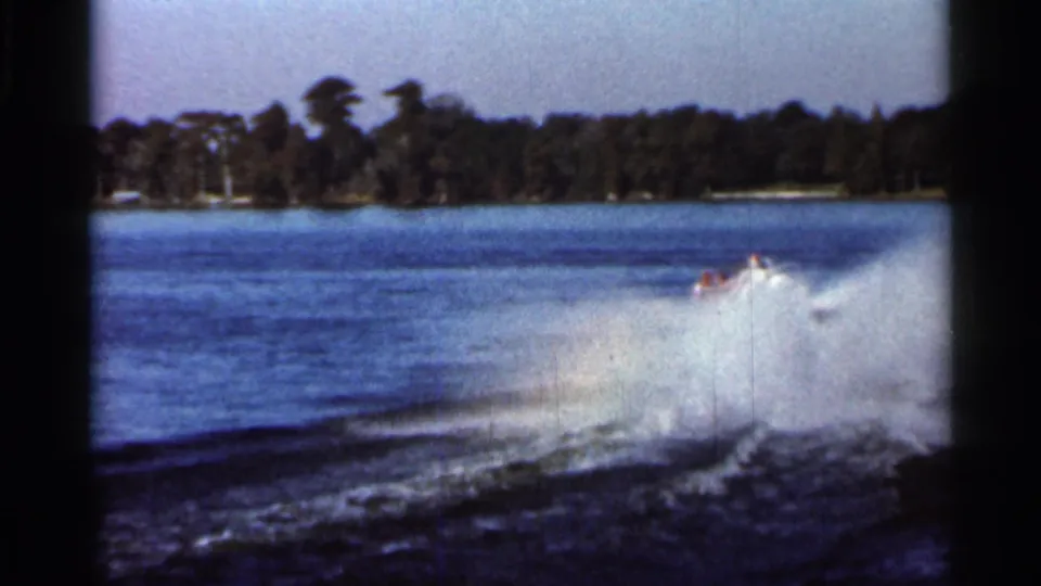 1966 Florida 8mm frame showing a partially obscured water skier or tow activity behind heavy spray on a lake, with shoreline trees and home-movie film borders visible.