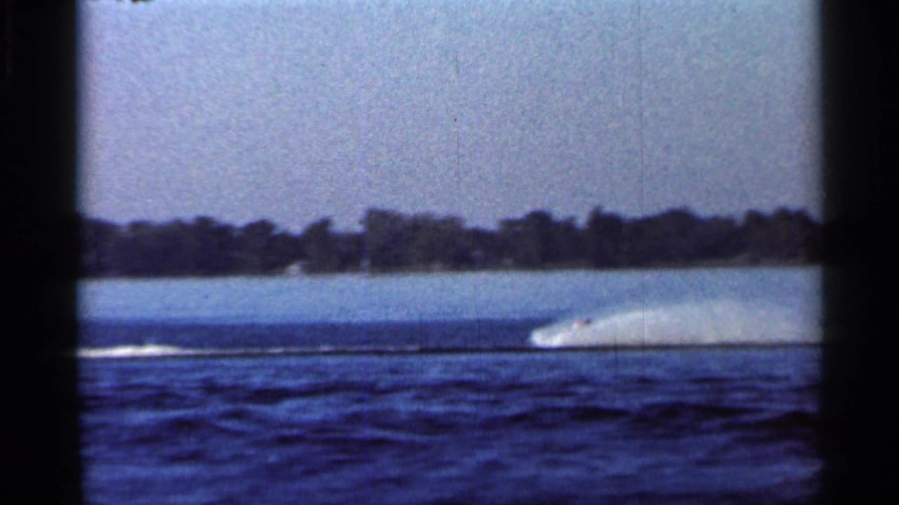 1966 Florida water ski show footage showing a distant speedboat throwing spray across a lake with wooded shoreline in the background.