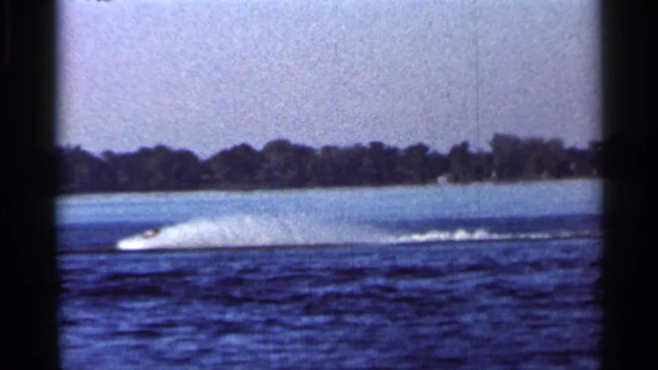 Distant boat during the Cypress Gardens water ski show, seen across blue lake water with wake and tree-lined shore.