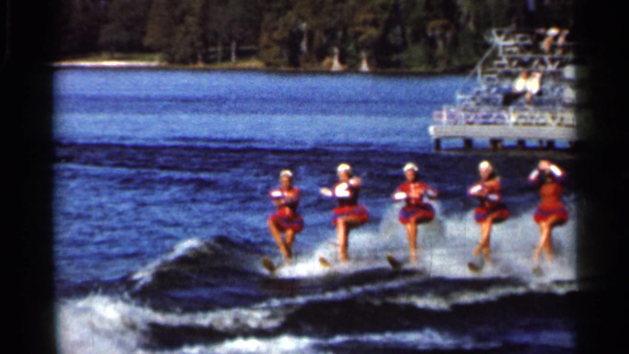 Four women water ski in a line across a lake with a dock and tree-lined shore behind them.