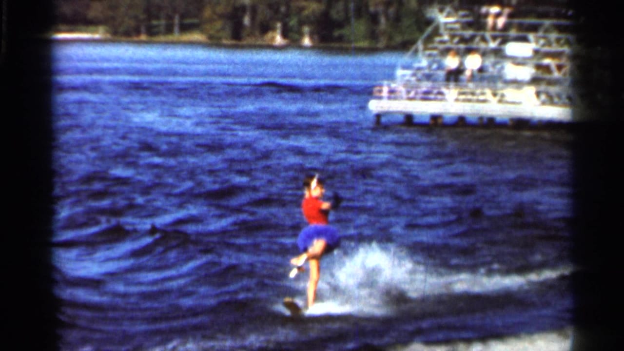 1966 Florida home-movie frame showing a woman in a red top and blue skirt water-skiing across a lake with a boat behind her.