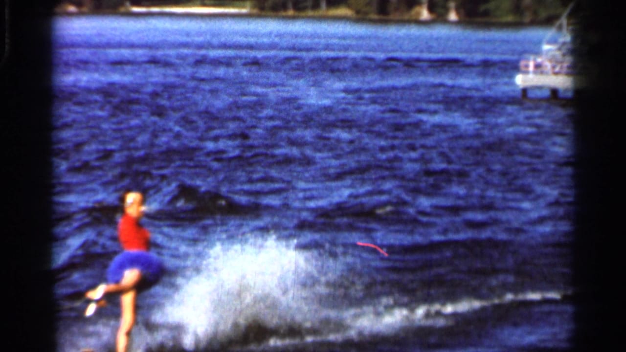 1966 Florida lake footage frame with the woman water-skier leaning through a turn as a spray plume rises behind her.