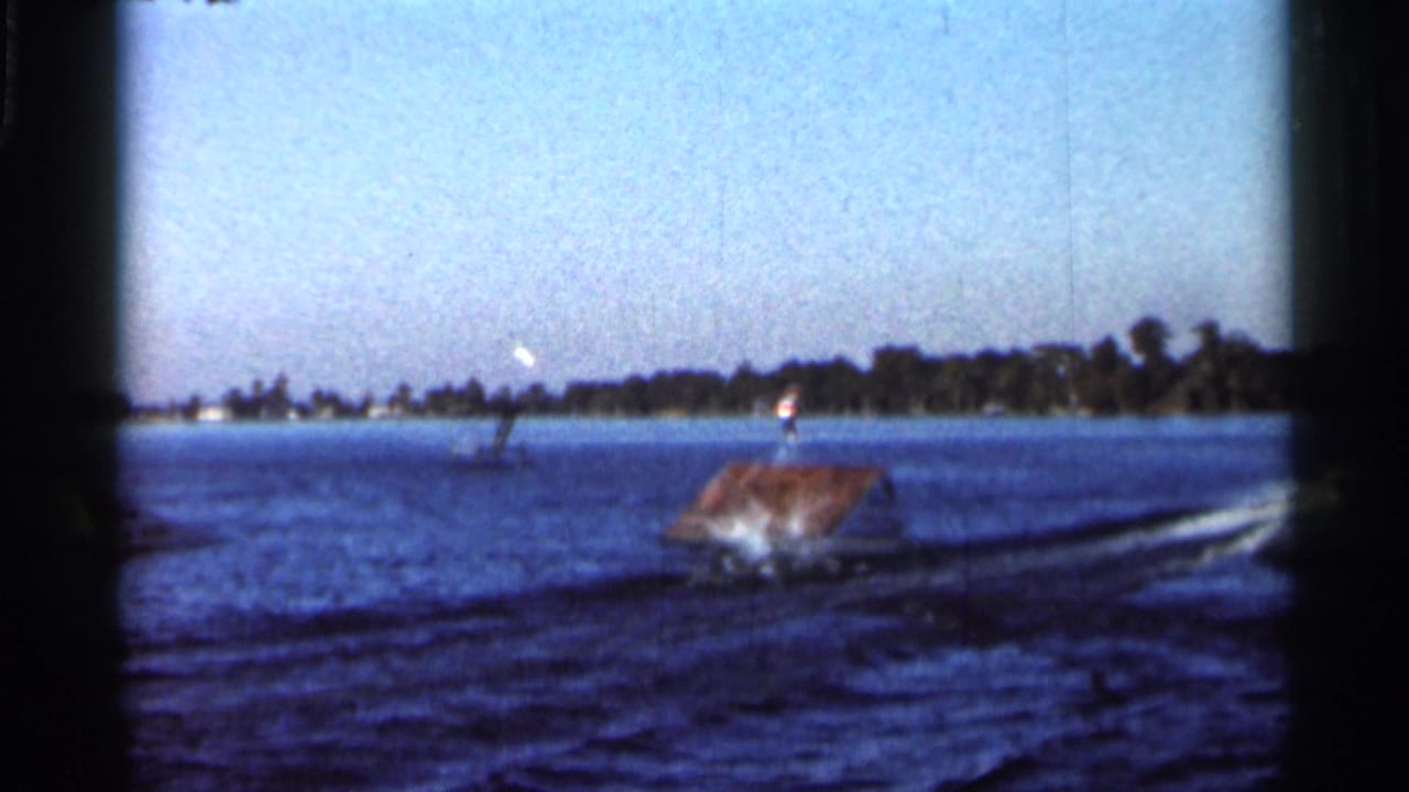 Wide 1966 Florida home-movie frame showing lake water, a small ramp or dock structure, a distant skier, shoreline, and skywriting in the sky.