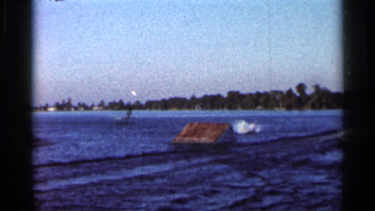 1966 Florida lake frame with a low ramp or dock, a distant water-skier, splash on the right, shoreline trees, and a skywriting trail.