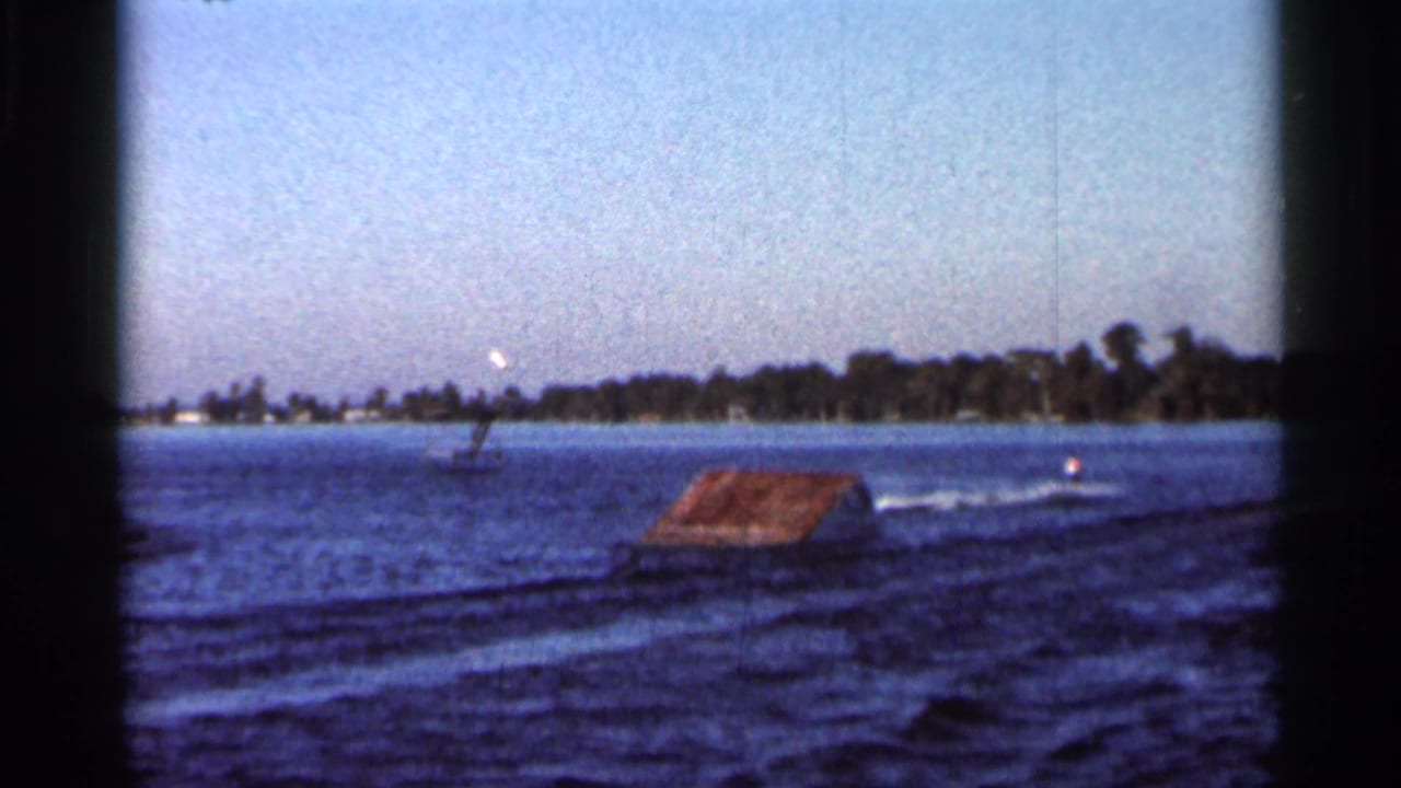 1966 Florida 8mm frame showing a wooden water ski ramp on a lake, with rippled water and a distant wooded shoreline.