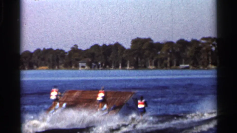 A distant water skier in a life vest approaches or hits a wooden jump ramp on a Florida lake, throwing spray behind them.