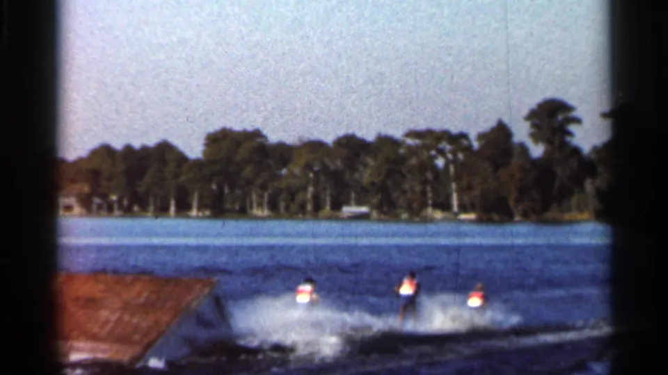 A water skier splashes down after the jump attempt, with the wooden ramp and wooded Florida shoreline visible behind the action.