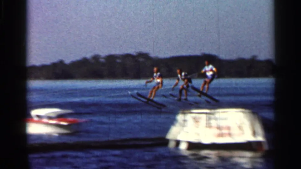 1966 Florida 8mm frame showing three visible water-sport participants skiing together near a boat and ramp on a lake.