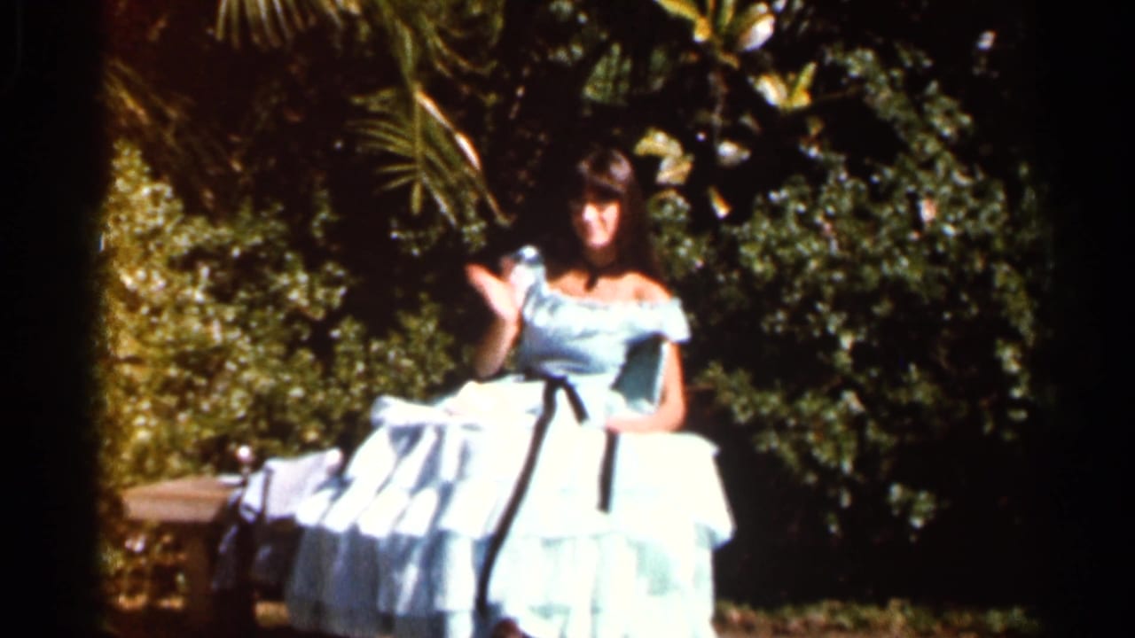 1966 Florida home-movie frame of a woman in a light blue off-the-shoulder gown posing among dense garden foliage.