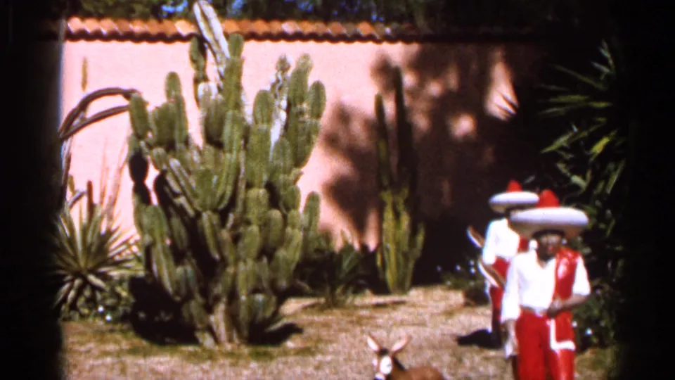 1966 Florida home-movie frame showing charro-style costumed figures near cactus, dry grass, and a sunlit stucco garden wall.
