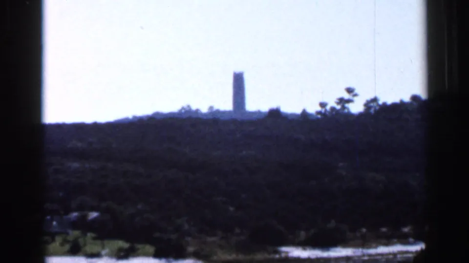 1966 Florida 8mm frame showing a tall distant tower silhouetted beyond a tree-covered hill under a pale bright sky.