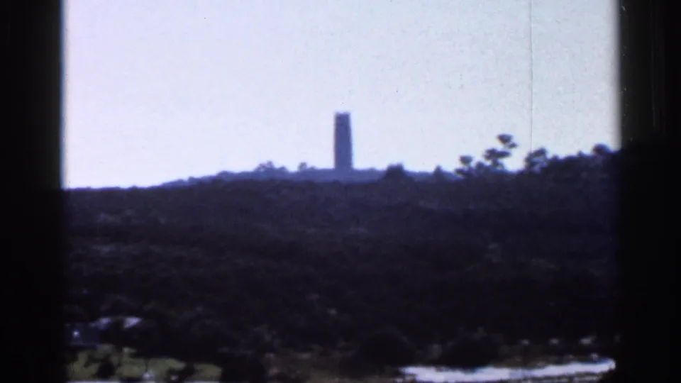 1966 Florida home-movie frame with a distant tower silhouette rising over a wooded hill and bright sky, framed by 8mm gate edges.