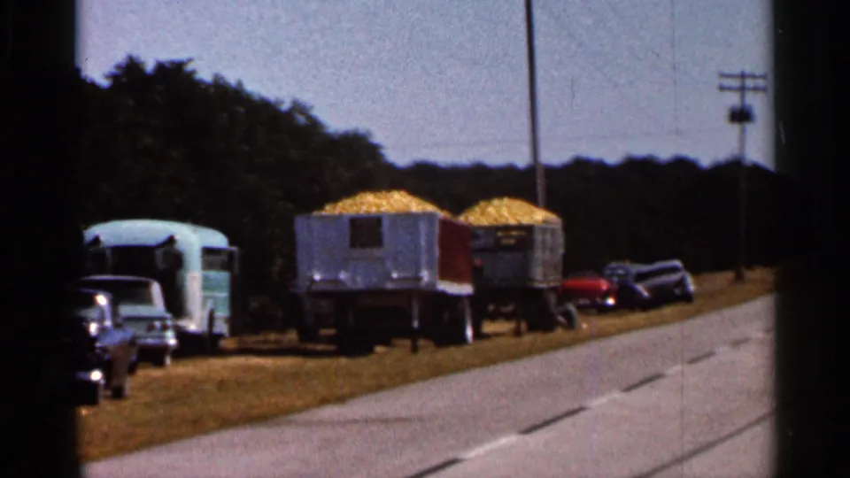 1966 Florida home-movie frame showing corn trailers, parked mid-century cars, a pale camper trailer, highway pavement, and trees.