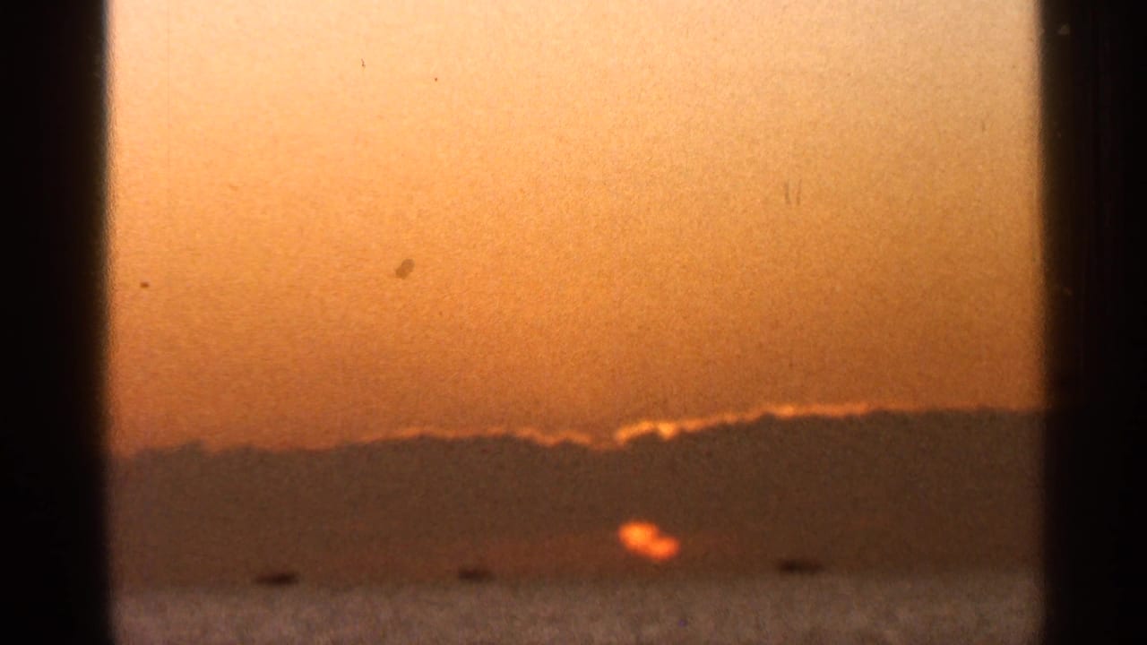 1966 Florida 8mm frame showing a low orange sun over water beneath a dark horizon, with visible film grain and small dust marks.
