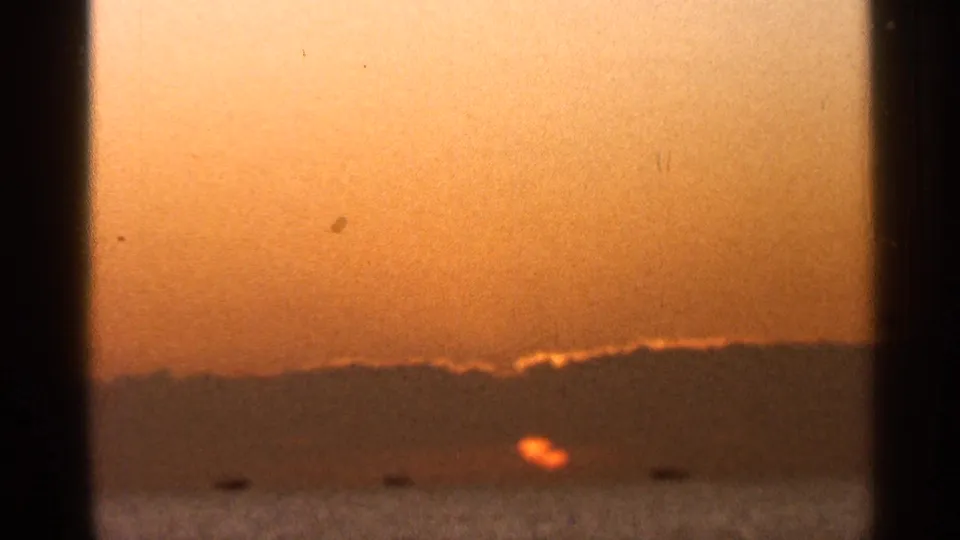 1966 Florida 8mm frame showing a low orange sun over water beneath a dark horizon, with visible film grain and small dust marks.