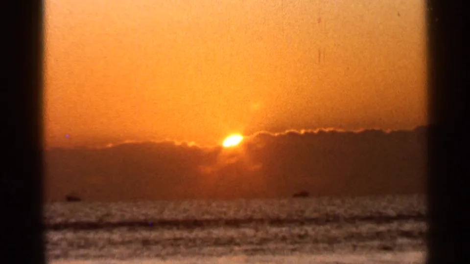1966 Florida 8mm frame of the sun dropping behind low clouds over the ocean, with warm haze and small dark shapes on the water.