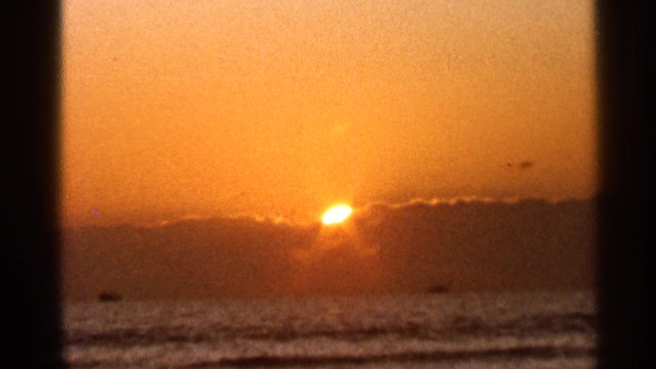 1966 Florida 8mm frame of a glowing sun at the cloud line above the ocean, with surf bands and a dark shoreline edge below.