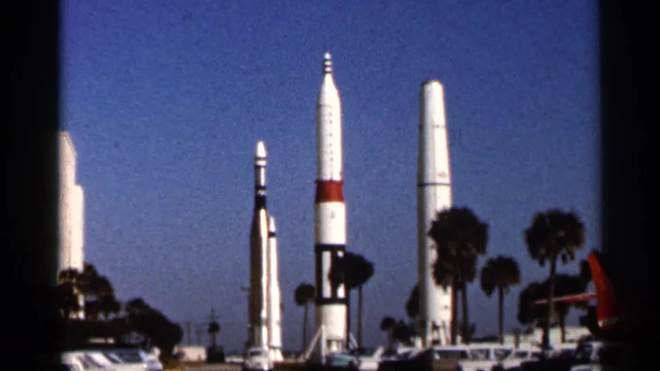 1966 Florida home-movie frame of several white rockets with red and black accents standing outdoors near palm trees and parked cars.