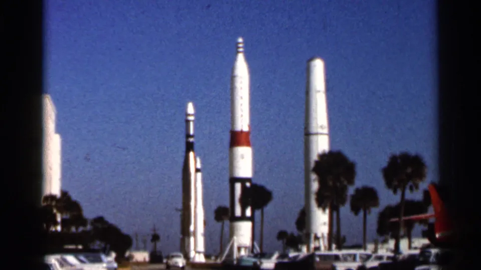 1966 Florida 8mm frame showing a visitor view of tall rocket displays with palm trees, cars, and strong sunlight.