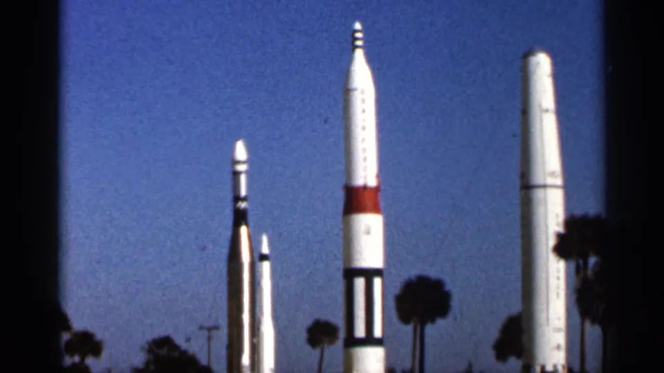 1966 Florida home-movie frame of white rocket displays with red and black markings, palm silhouettes, and a clear blue sky.
