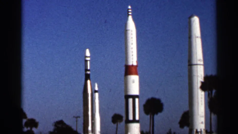 1966 Florida home-movie frame with a lineup of white rockets, dark banding, and palm trees at an outdoor space exhibit.