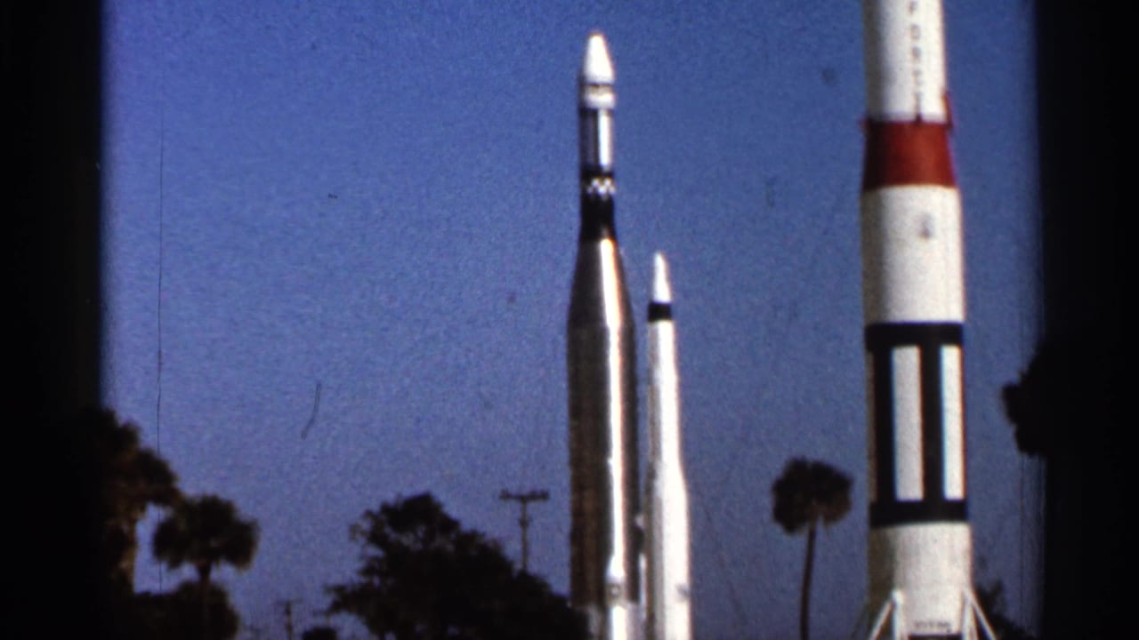 1966 Florida home-movie frame showing three rocket-like structures outdoors, with palm trees and deep blue sky in the background.