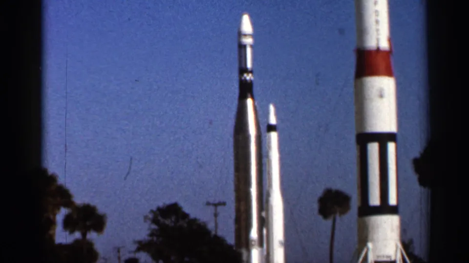 1966 Florida home-movie frame showing three rocket-like structures outdoors, with palm trees and deep blue sky in the background.