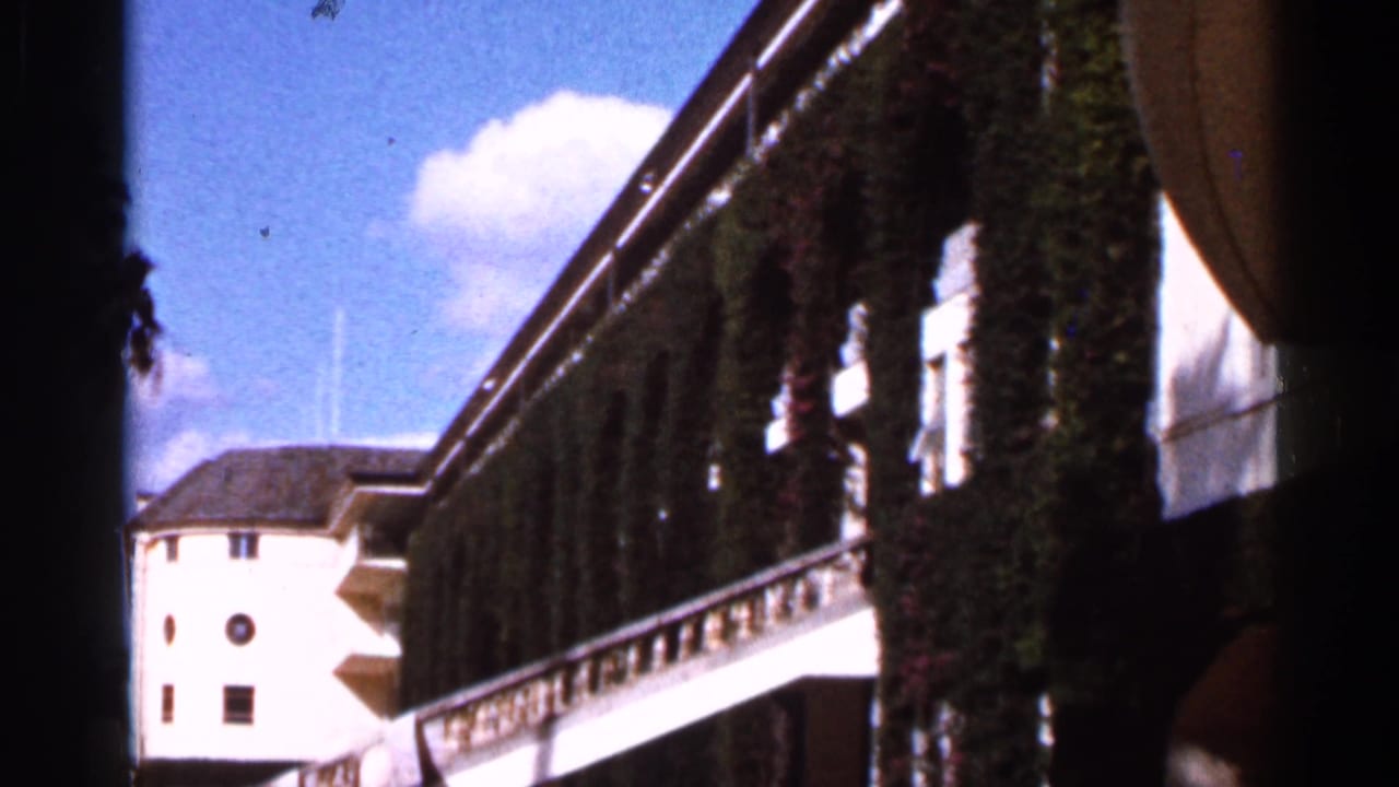 1966 Florida 8mm frame showing a white building with brown trim, ivy-covered sections, balcony railing, blue sky, and clouds.