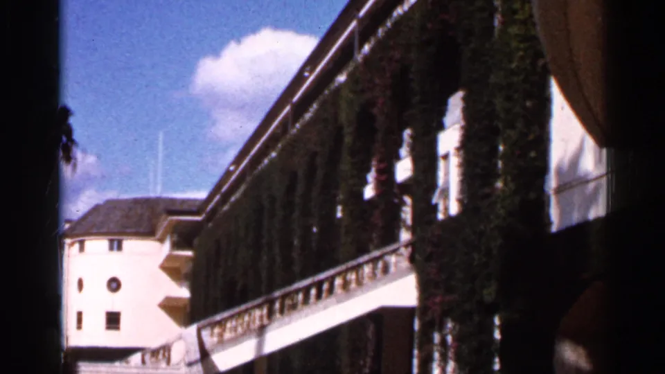 1966 Florida 8mm frame of a white building facade with ivy, balcony railing, brown trim, and blue sky in strong daylight.