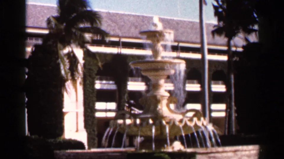 1966 Florida home-movie frame showing a three-tiered fountain, palm trunks, and a building with arched openings behind it.