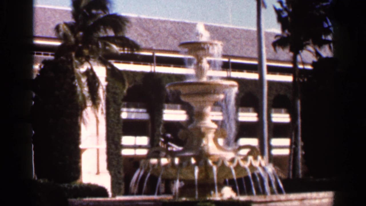 Tiered fountain spraying water in front of palms and an arched building facade.
