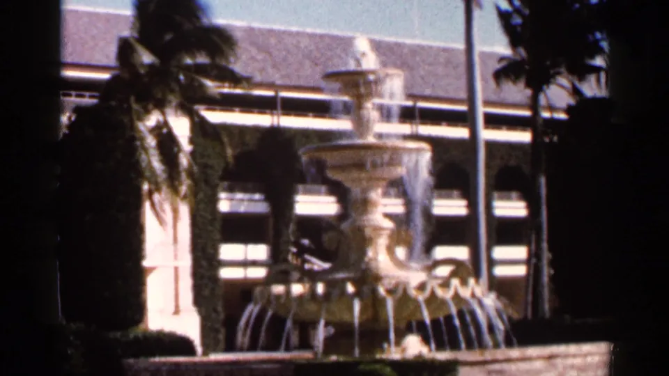 A vintage 8mm Florida view holds on a tiered outdoor fountain, palm foliage, and a building facade with arched windows.