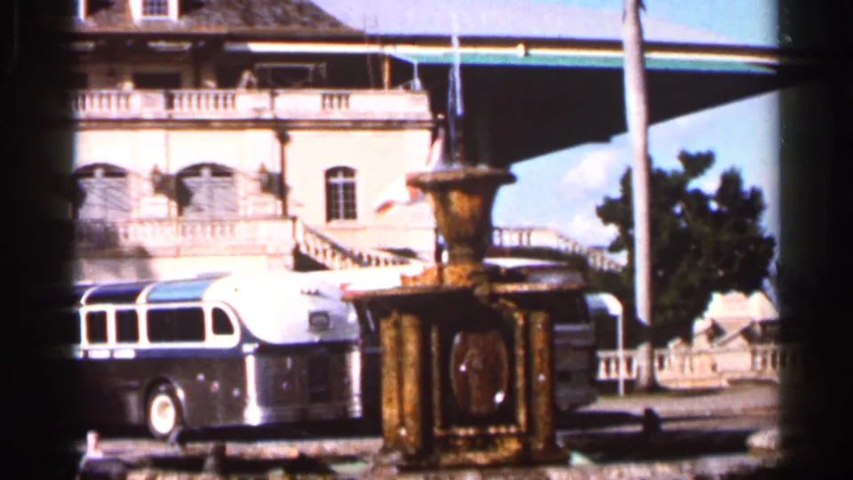 1966 Florida 8mm frame showing a fountain, parked mid-century bus, and ornate building facade with balconies and arched windows in sun.