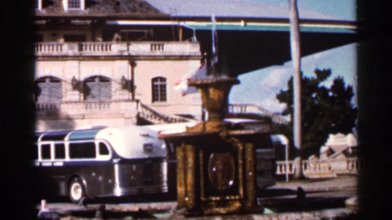 Ornate fountain beside a silver-and-white bus in front of a large balcony-fronted building.