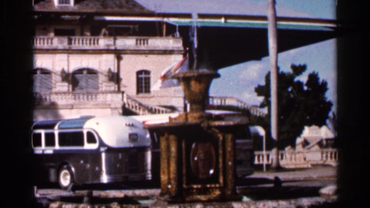 Classic bus parked near an ornate fountain with a large building and trees behind it.
