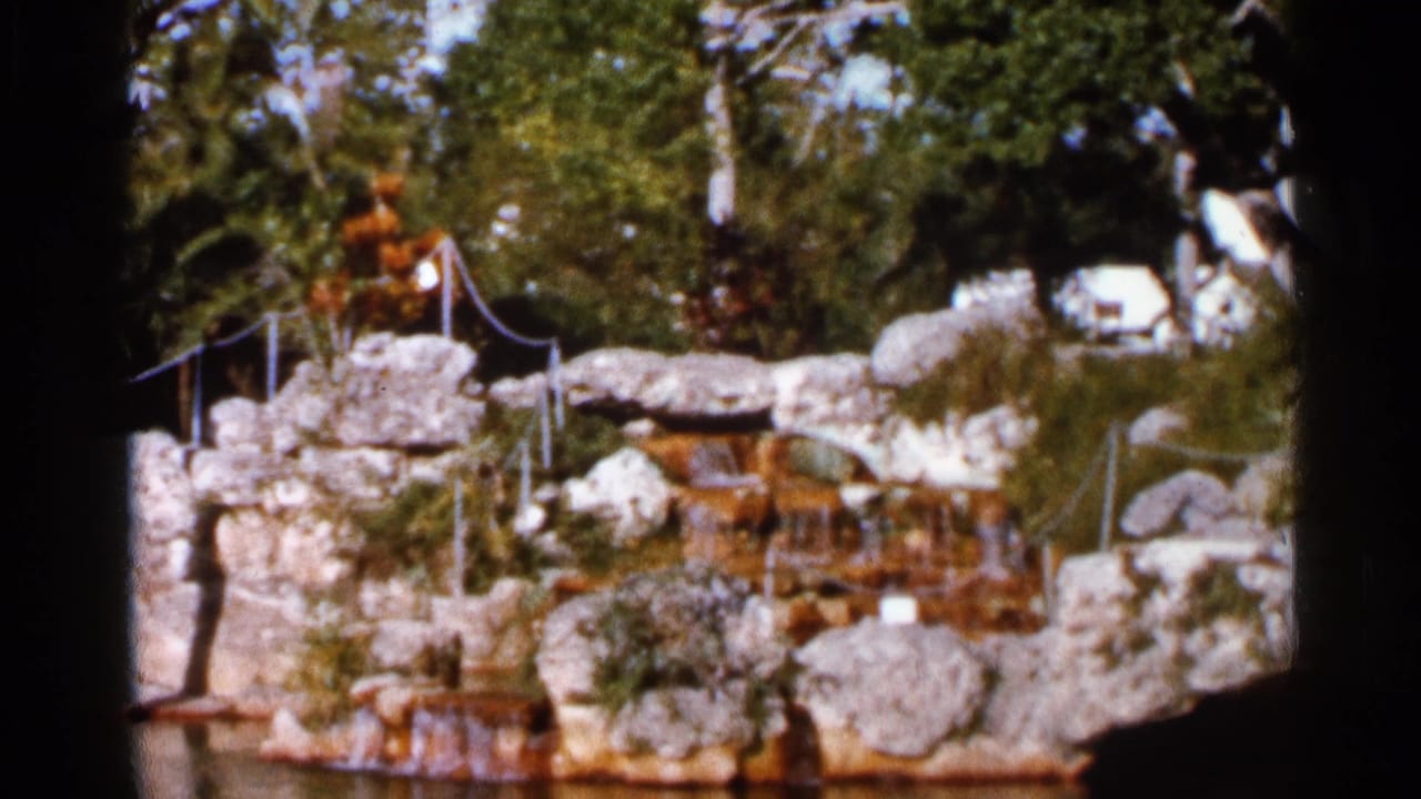 Rock waterfall with rope railing, trees, shrubs, and a small pool in bright sun.