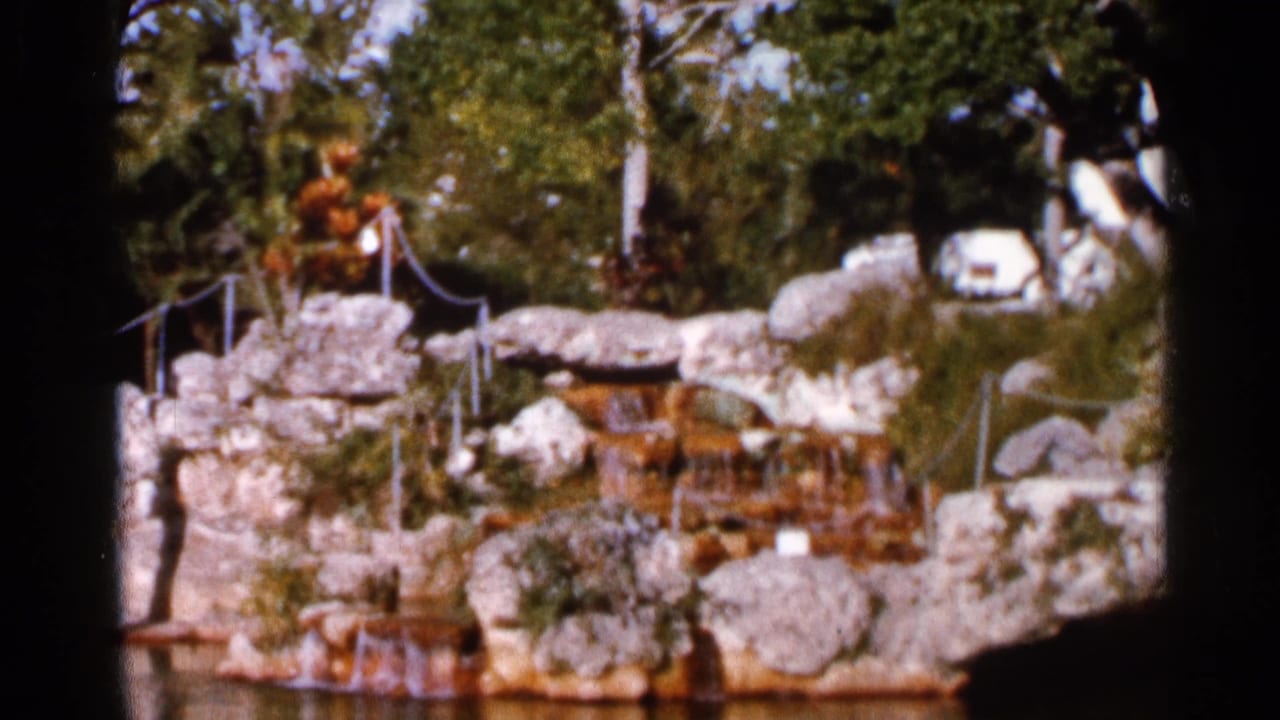 1966 Florida home-movie frame of a sunlit rock waterfall feature with rope railings, trees, foliage, and water below.