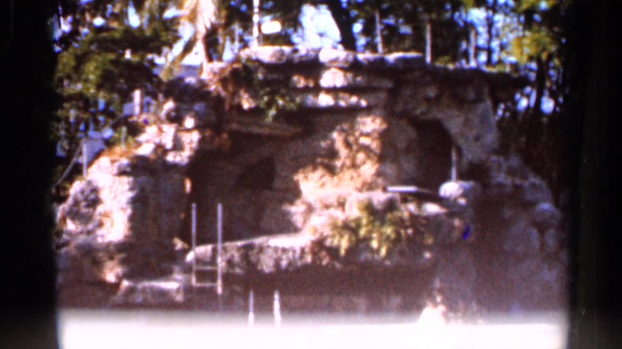 1966 Florida 8mm frame showing the lower pool and rock wall of a landscaped waterfall area, with railings and tree shade.