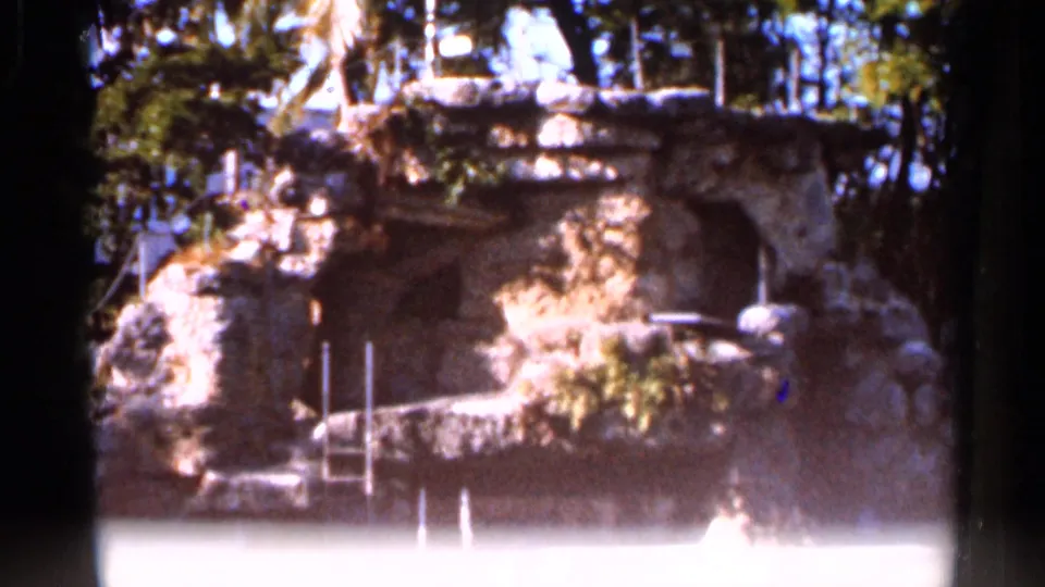 1966 Florida 8mm frame showing the lower pool and rock wall of a landscaped waterfall area, with railings and tree shade.