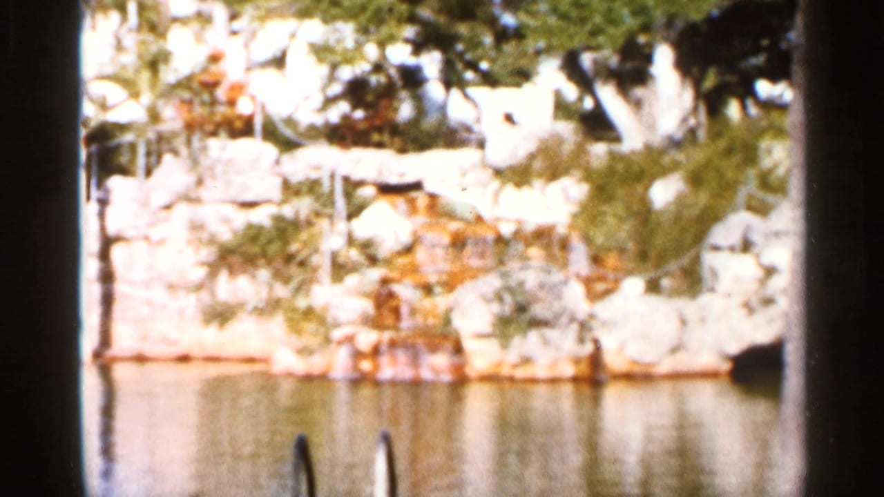 Sunlit rock wall with small waterfall beside calm water and ladder rails.