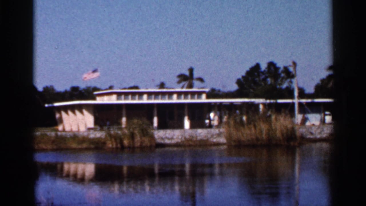 Low mid-century building beside a Florida waterway, with an American flag above and reeds along the bank.