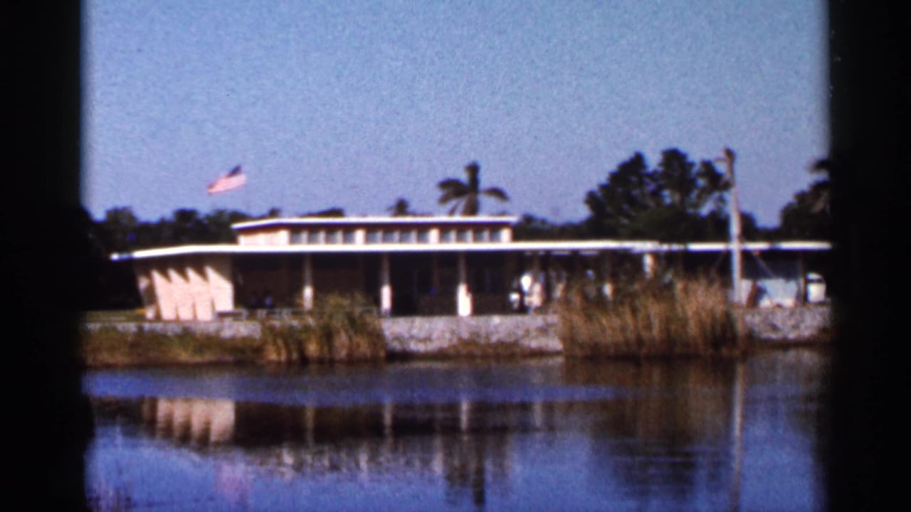 1966 Florida home-movie view of a low building reflected in water, with reeds, trees, palms, and a flag visible.