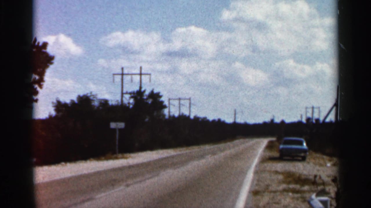 1966 Florida home-movie frame showing a highway stretching ahead under scattered clouds, with a lone car at right and power lines along the roadside.