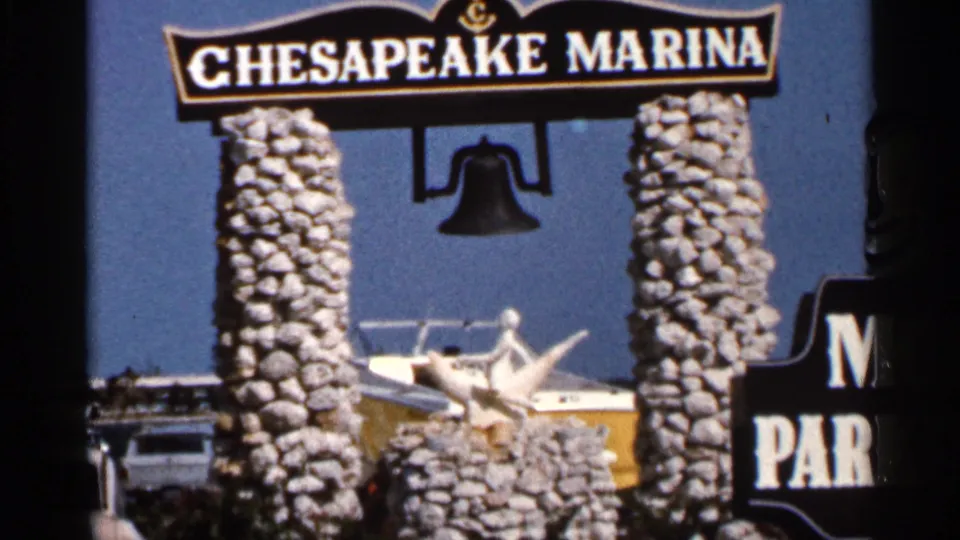 1966 Florida 8mm frame showing the Chesapeake Marina entrance sign, stone supports, hanging bell, boat, and partial roadside parking sign.