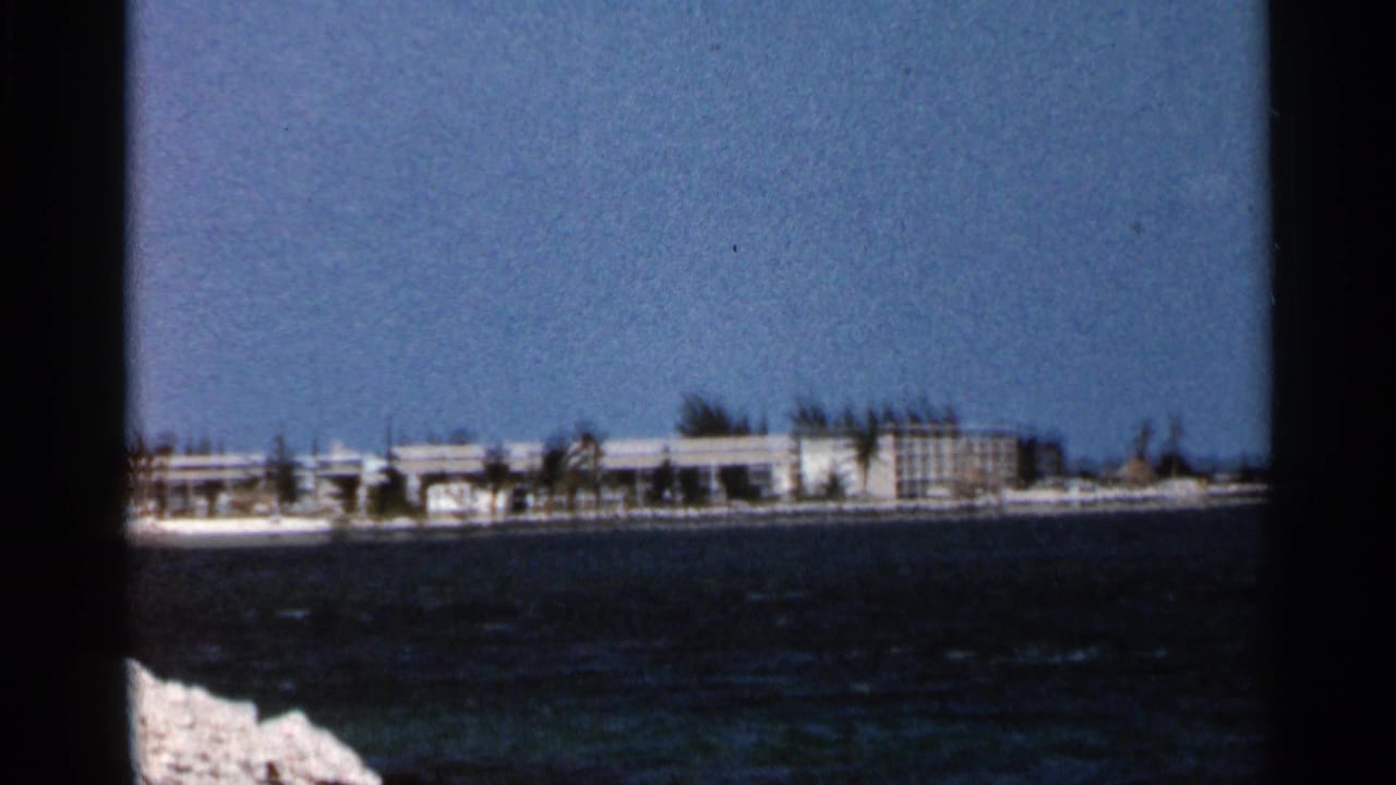 1966 Florida home-movie view of a low coastal resort complex seen across choppy blue water, with pale rocks in the foreground.