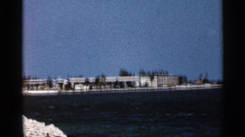 1966 Florida home-movie view of a low coastal resort complex seen across choppy blue water, with pale rocks in the foreground.