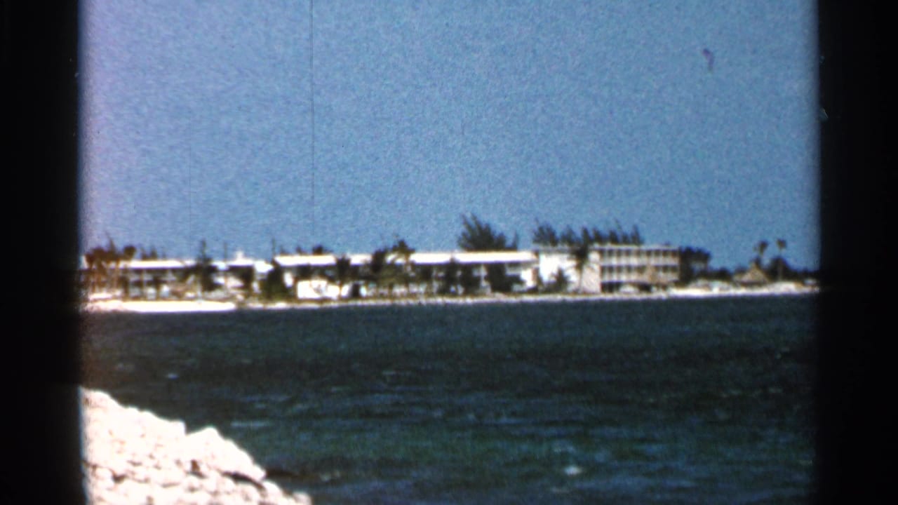 1966 Florida shoreline view with low resort buildings, palm trees, a sandy strip, and deep blue water in the foreground.
