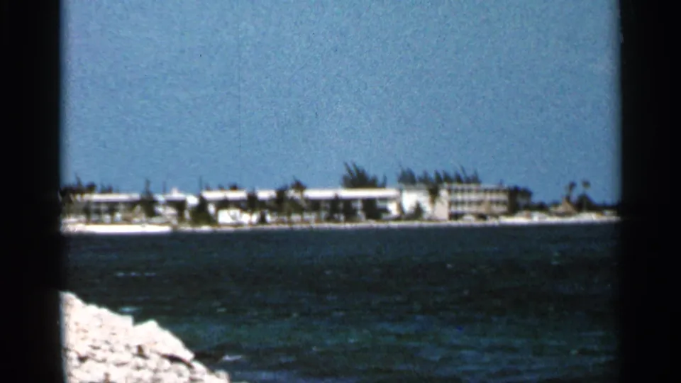1966 Florida home-movie frame showing a resort shoreline across rippled water, with palms and pale rocks at the edge of the frame.