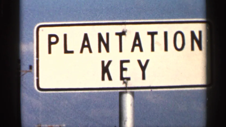 1966 Florida frame with a white road sign reading Plantation Key in black letters against a clear sky.
