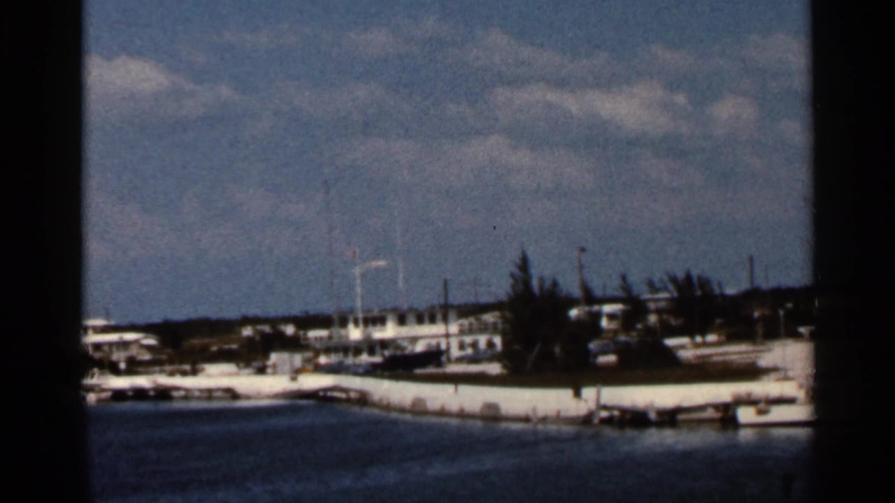 Distant waterfront with boats, white buildings, trees, and blue water under a cloudy sky.